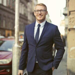 Smiling businessman in suit with briefcase walking on urban street with parked cars.