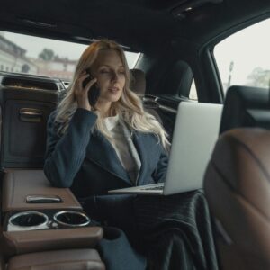 Confident businesswoman making a call and working on a laptop in a luxury car interior.